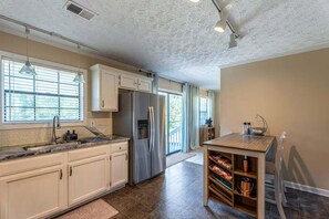 A modern kitchen with a stainless steel refrigerator, white cabinets, a built-in oven, and a dining area with a wooden table and bookshelves. - Peachtree City Home Near Lake with Yard & Firepit (Peachtree City)