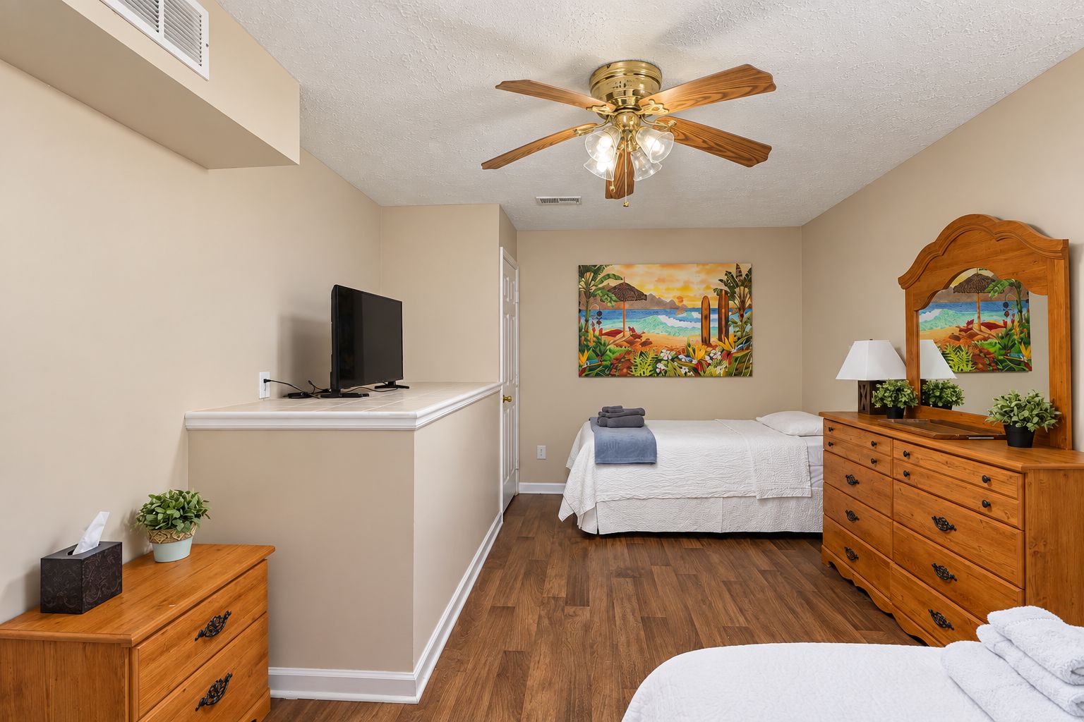 A bedroom with a bed, a TV on a stand, a ceiling fan, a painting, a dresser, and a mirror.