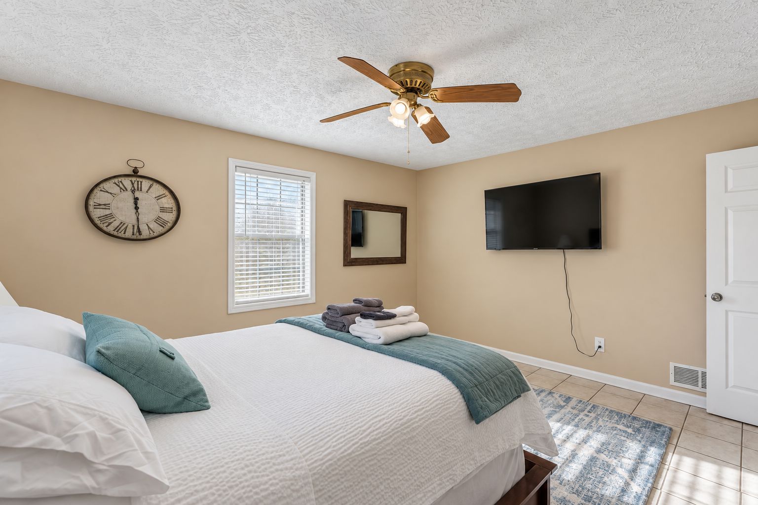 Une chambre à coucher avec un lit, un ventilateur de plafond, une horloge, une fenêtre, un téléviseur et une porte.
