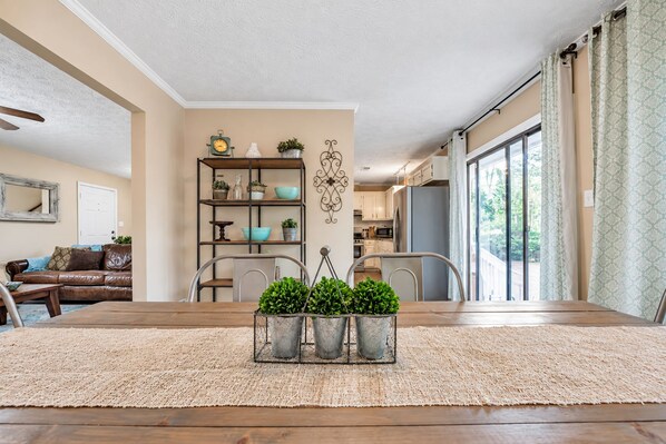 A dining area with a wooden table, chairs, and a woven rug. There is a kitchen in the background with white cabinets and a large window.