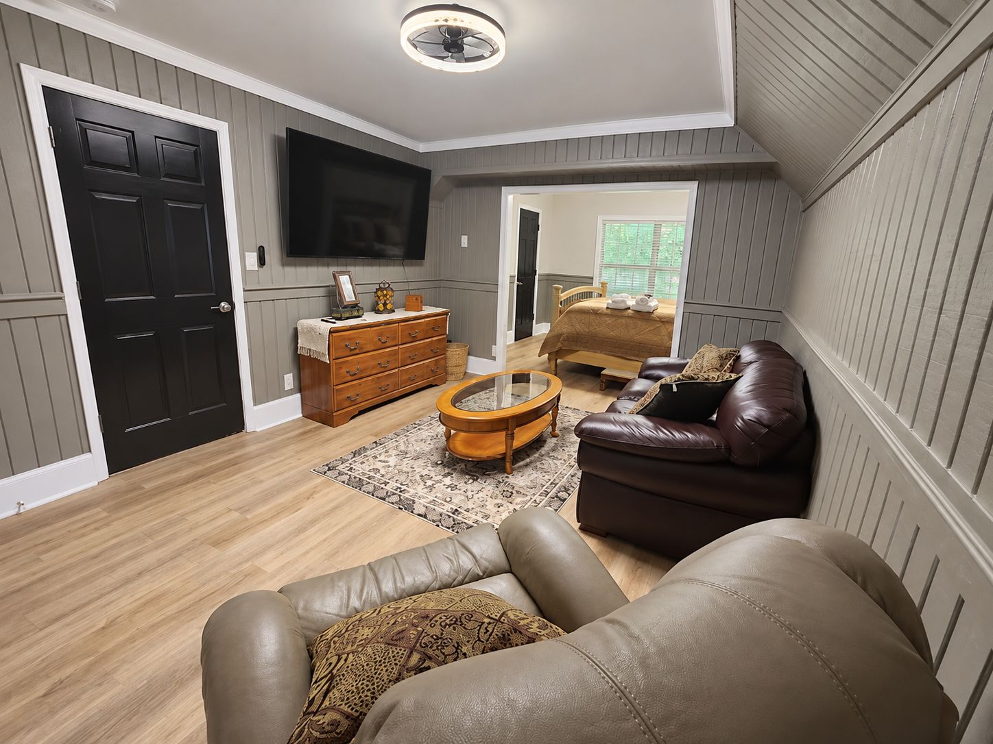 A living room with a leather sofa, a wooden coffee table, a TV mounted on the wall, and a bed with a wooden headboard in the background.