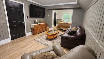 A living room with a leather sofa, a wooden coffee table, a TV mounted on the wall, and a bed with a wooden headboard in the background.