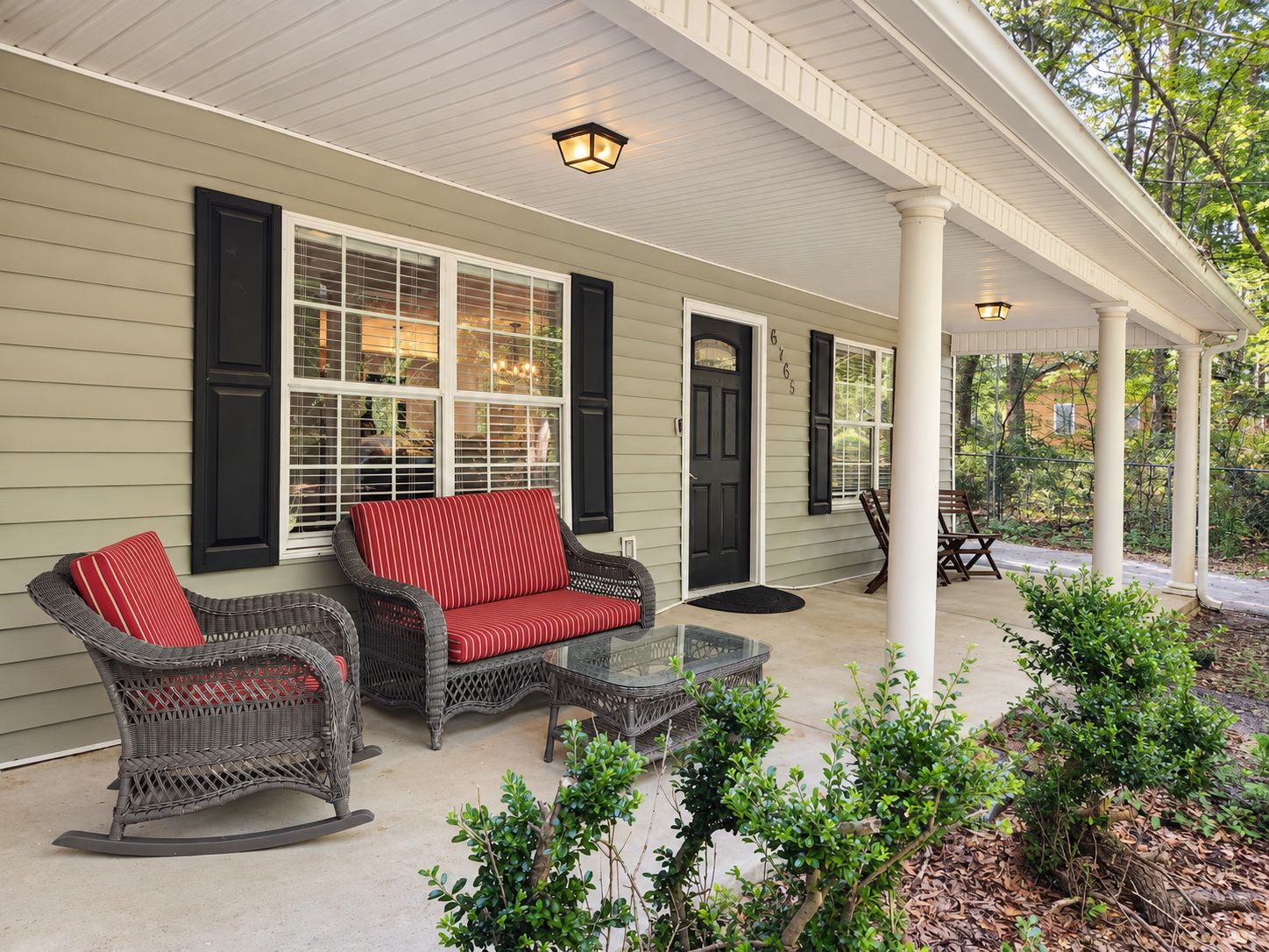 A covered porch with wicker furniture, a glass table, and a black door.