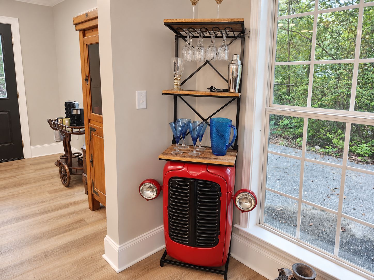 A vintage red refrigerator with a shelf holding glasses and a bottle, a wooden cart with kitchen items, and a window with a view of greenery.