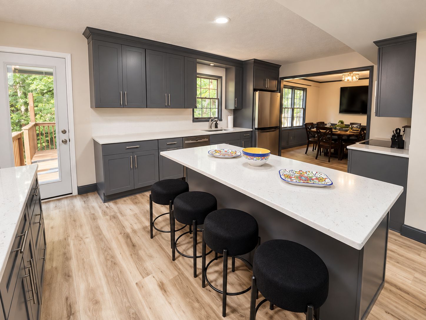 A modern kitchen with dark cabinets, a central island with stools, and a dining area visible through an open door.