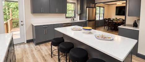 A modern kitchen with dark cabinets, a central island with stools, and a dining area visible through an open door.