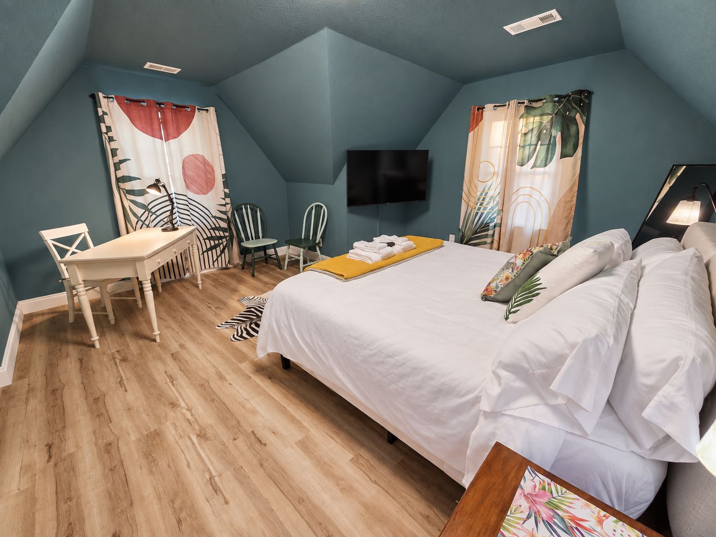 A bedroom with a bed, a desk, two chairs, a flat-screen TV, and a zebra-patterned rug.