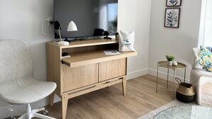 Ein modernes Wohnzimmer mit einem Holztisch, einem Flachbildfernseher, einem weißen Sessel, einem kleinen Beistelltisch mit Pflanze und einem gerahmten Blumenbild an der Wand.