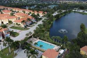 A residential area with a swimming pool, houses with red roofs, and a lake with a fountain. - Relaxed Florida Living | Gated Community with Heated Pool | Near Beaches, Shopping & Dining (Fort Myers)