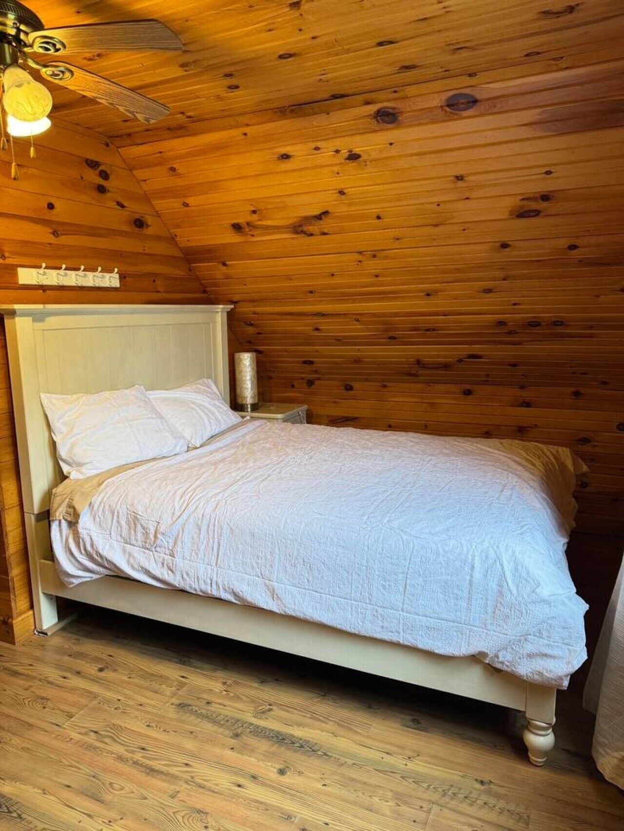 A single bed with a white headboard and a bedside table with a lamp in a room with wooden walls and a sloped ceiling.