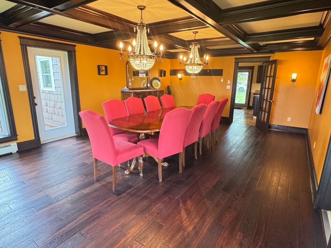 A dining area with a wooden table and red chairs, chandeliers, and a view through a glass door.