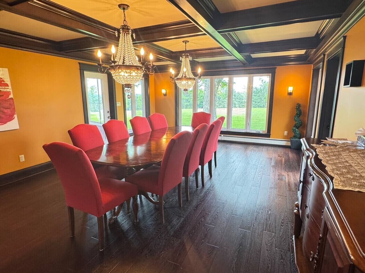 Un comedor con mesa de madera, sillas rojas, ventanas amplias y una araña.