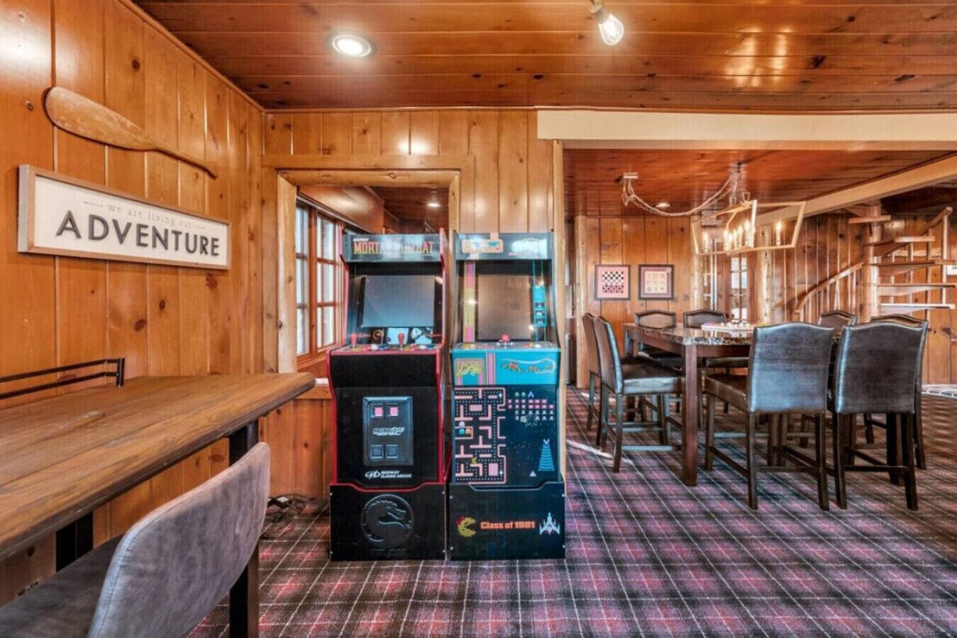 Un intérieur en bois avec une enseigne indiquant « AVENTURE », une borne d’arcade rétro, un coin repas avec des chaises et une table, et un sol à motifs écossais.