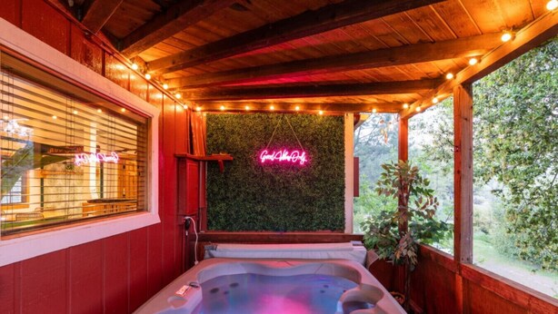 A wooden interior with a hot tub, a neon sign, and a view of greenery. - River Falls - Hot Tub and Mountain Views sleeps 4 (Oakhurst)
