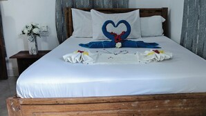 A neatly made bed with a heart-shaped towel arrangement, a wooden headboard, and a nightstand with a vase of flowers. - Primavera Jambiani (Jambiani)