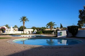 A swimming pool surrounded by white buildings with palm trees, a clear sky, and a paved area. - Algarve escape in the heart of the prestigious Golden Triangle (Quarteira)