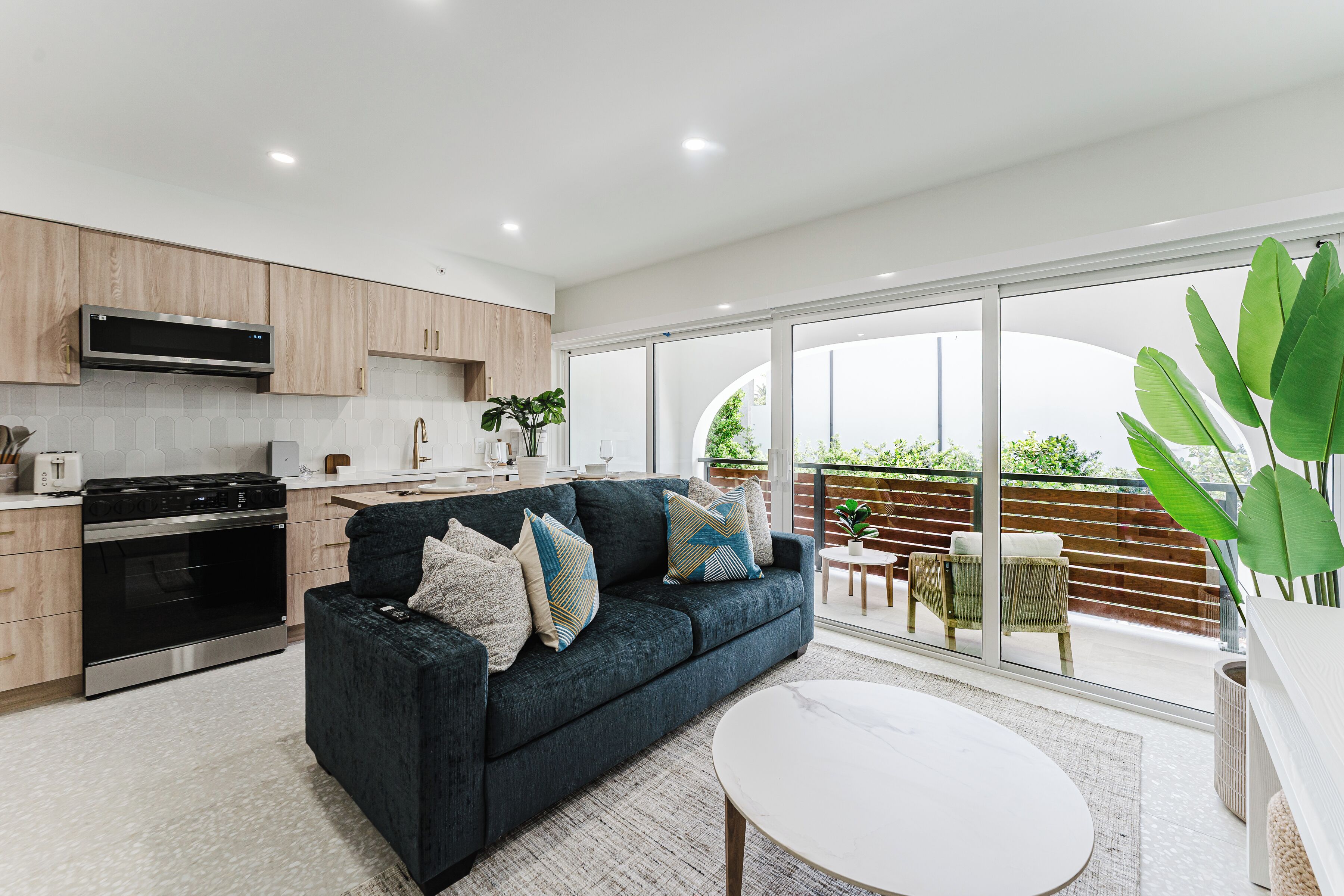 A modern kitchen with built-in appliances, a dark-colored sofa with decorative pillows, and a large sliding glass door leading to an outdoor area.
