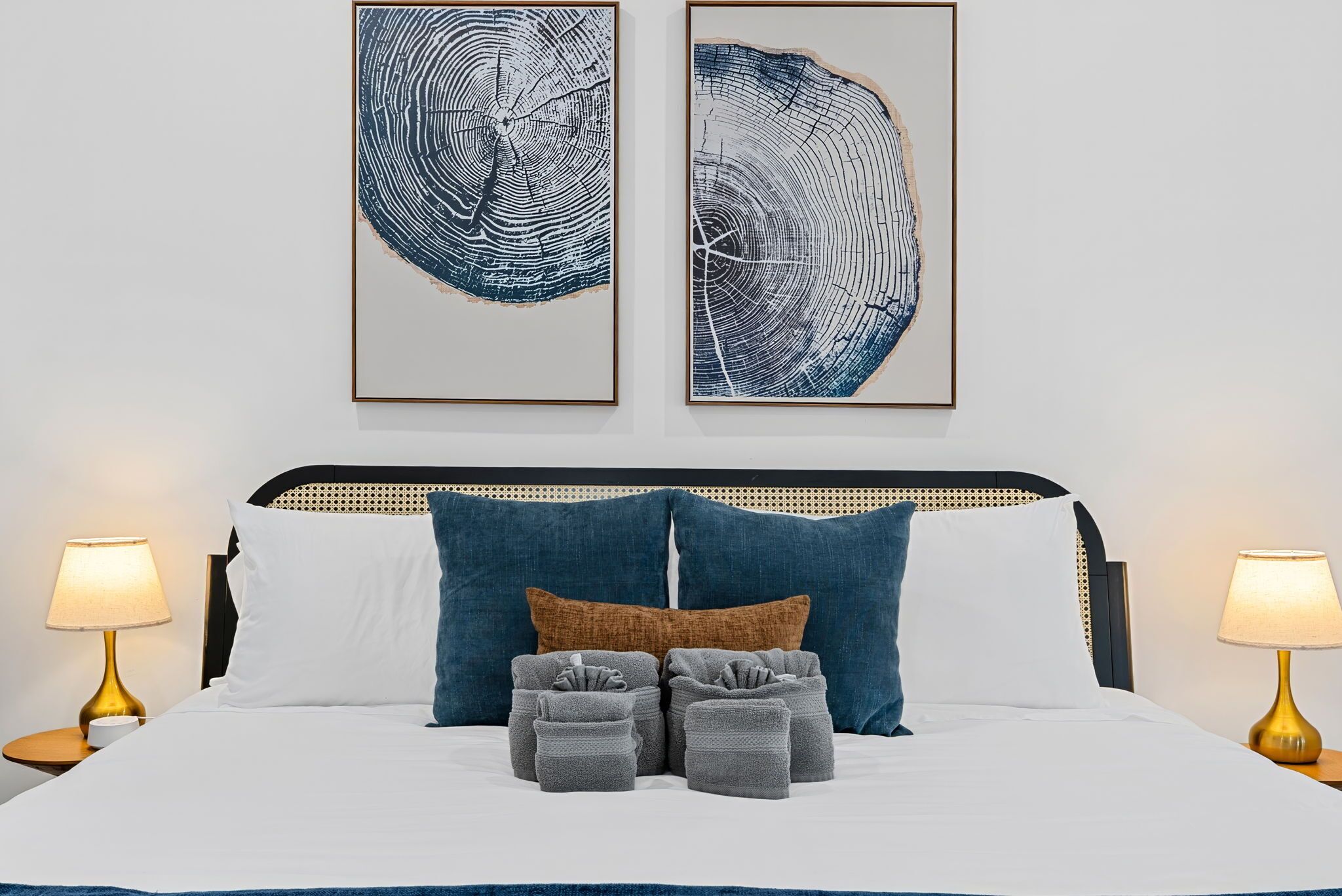 A bed with white bedding, two blue pillows, a brown pillow, and three gray throw pillows. There are two bedside lamps with gold bases. Two framed artworks are hung above the bed.