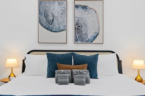 Ein Bett mit weißer Bettwäsche, zwei blaue Kissen, ein braunes Kissen und drei graue Zierkissen. Zwei Nachttische mit goldfarbenen Füßen stehen daneben. Darüber hängen zwei gerahmte Kunstwerke.