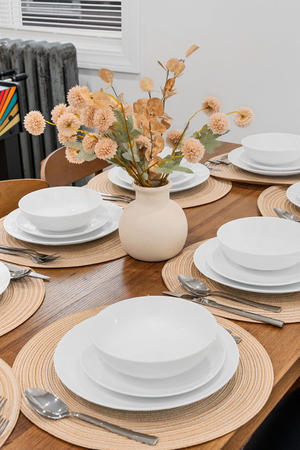 A dining table set with white plates, silverware, and a vase with dried flowers.