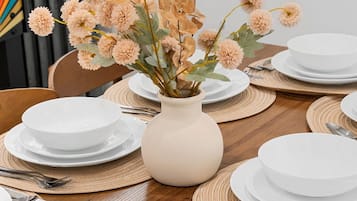 A dining table set with white plates, silverware, and a vase with dried flowers.