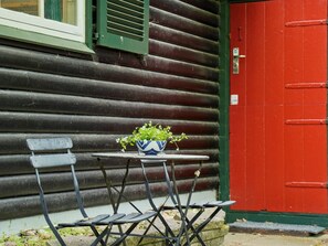 Eine Holzhütte mit roter Tür, einem kleinen Tisch mit Blumentopf und zwei Metallstühlen.