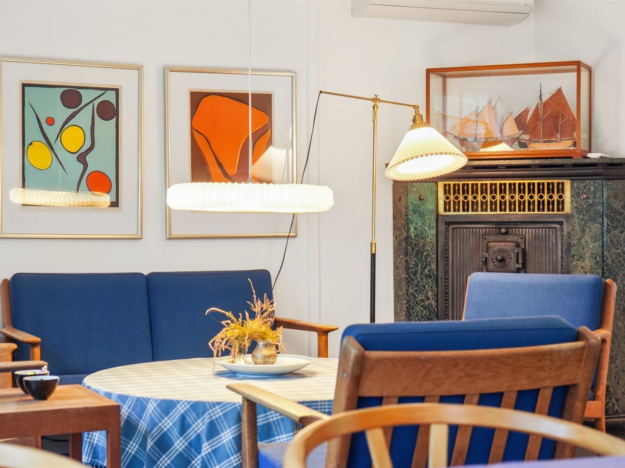 Een kamer met een blauwe bank, een ronde tafel met een geruit tafelkleed, een lamp en twee kunstwerken in lijsten aan de muur.
