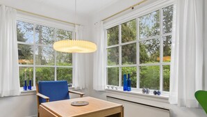 Huoneessa on puinen pöytä, siniset ja vihreät tuolit, riippuvalaisin ja suuret ikkunat, joissa on valkoiset verhot.