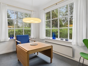 Ein Zimmer mit Holztisch, blauen und grünen Stühlen, einer Hängelampe und großen Fenstern mit weißen Vorhängen.