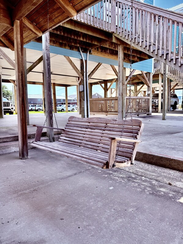 A wooden bench under a covered area with a metal railing, concrete floor, and a view of parked vehicles and buildings in the background.