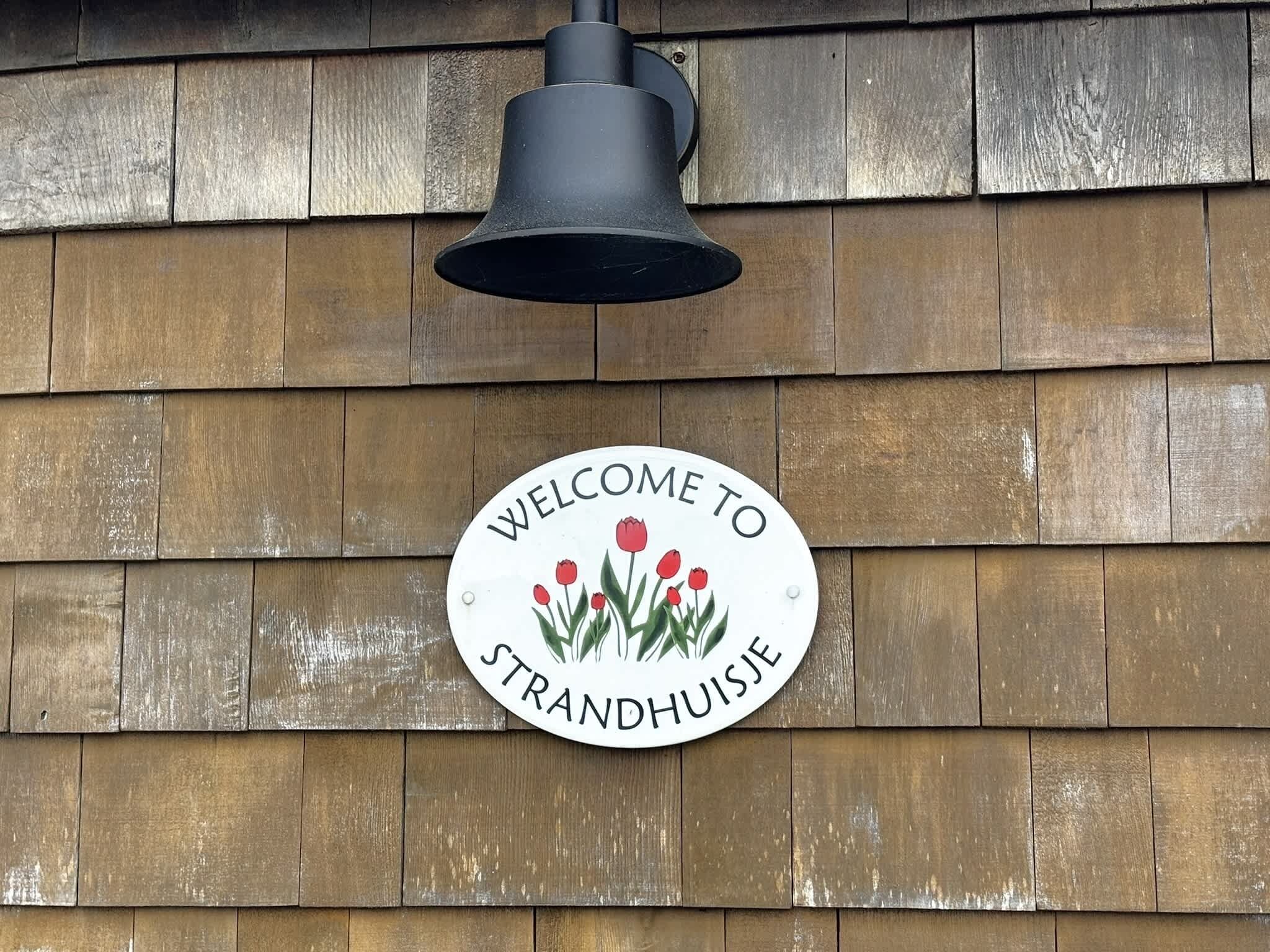 Une pancarte d’accueil avec l’inscription « BIENVENUE À STRANDHUISJE » et une cloche au-dessus.