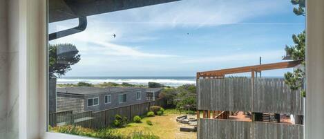 A view from a window overlooking a wooden deck, a grassy yard, and a beach with the ocean in the background.