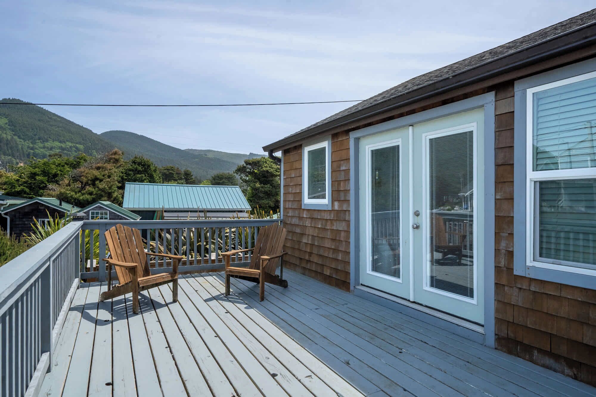 A wooden deck with two chairs, a view of mountains, and a house with large windows.