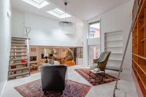 Un vaste espace de vie doté d’un haut plafond, de fenêtres de toit, d’un canapé en cuir noir, d’un tapis à motifs et d’une chaise en bois.