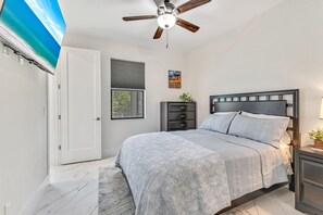 Ein Schlafzimmer mit einem Bett, einem Deckenventilator, einem Fenster mit Jalousien, einem Nachttisch und einem an der Wand befestigten Fernseher.