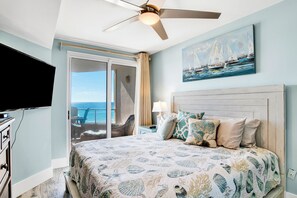 A bedroom with a bed, a ceiling fan, a TV, a painting of sailboats, and a view of the ocean through a sliding door.
