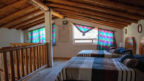 Une chambre avec deux lits, des poutres apparentes en bois au plafond, une fenêtre agrémentée de rideaux colorés et une balustrade en bois.