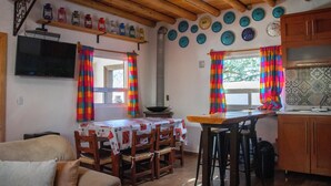 Une cuisine avec des meubles en bois, une table à manger avec des chaises, des rideaux colorés et un téléviseur fixé au mur.
