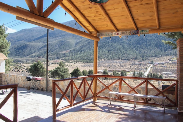 A covered patio with a view of a mountainous landscape, a wooden railing, and a table.