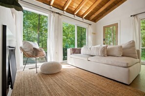 A modern living room with a white sofa, a round ottoman, and a chair with a cushion. There are large windows with curtains, a wooden ceiling, and a view of trees outside. - Forest Cottage w Fire Pit Near Cashiers! (Cashiers)