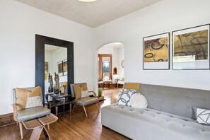 Ein modernes Wohnzimmer mit einem grauen Sofa, zwei Sesseln, einem Couchtisch und einem großen Spiegel. An den Wänden befinden sich abstrakte Gemälde und der Boden ist mit Holz ausgelegt.