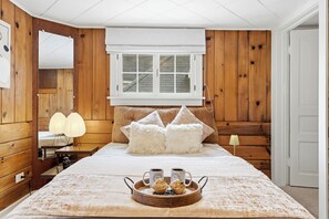 Ein Schlafzimmer mit einer holzverkleideten Wand, einem großen Bett mit flauschigen Kissen, einem Nachttisch mit Lampe sowie einem Tablett mit Muffins und Kaffeetassen.