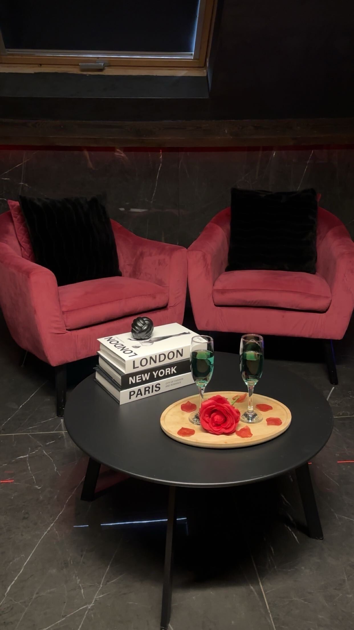 A room with two red sofas, a round black table with a rose and glasses, and a bookshelf with books titled 'LONDON,' 'NEW YORK,' and 'PARIS.'
