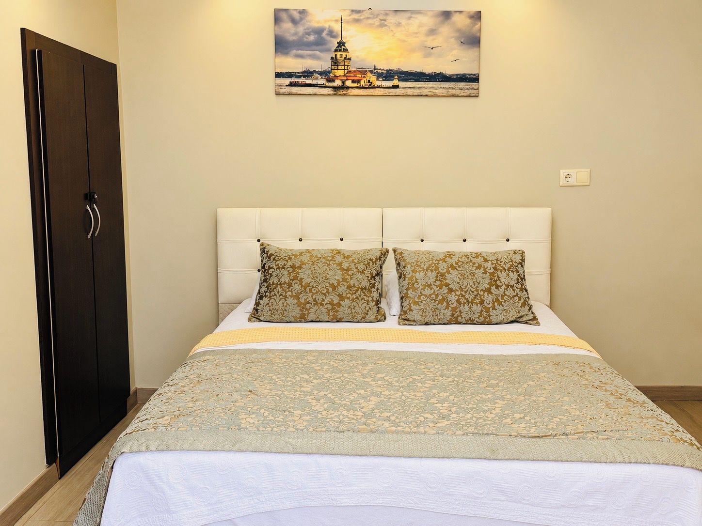 A bedroom with a bed, two pillows, a headboard, a painting on the wall, and a wardrobe.