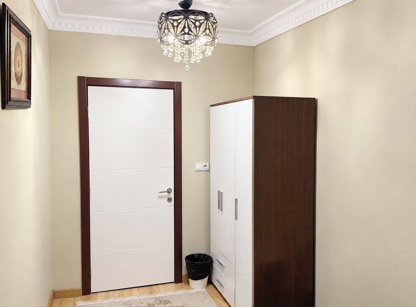 A hallway with a white door, a wooden wardrobe, a trash bin, and a chandelier.