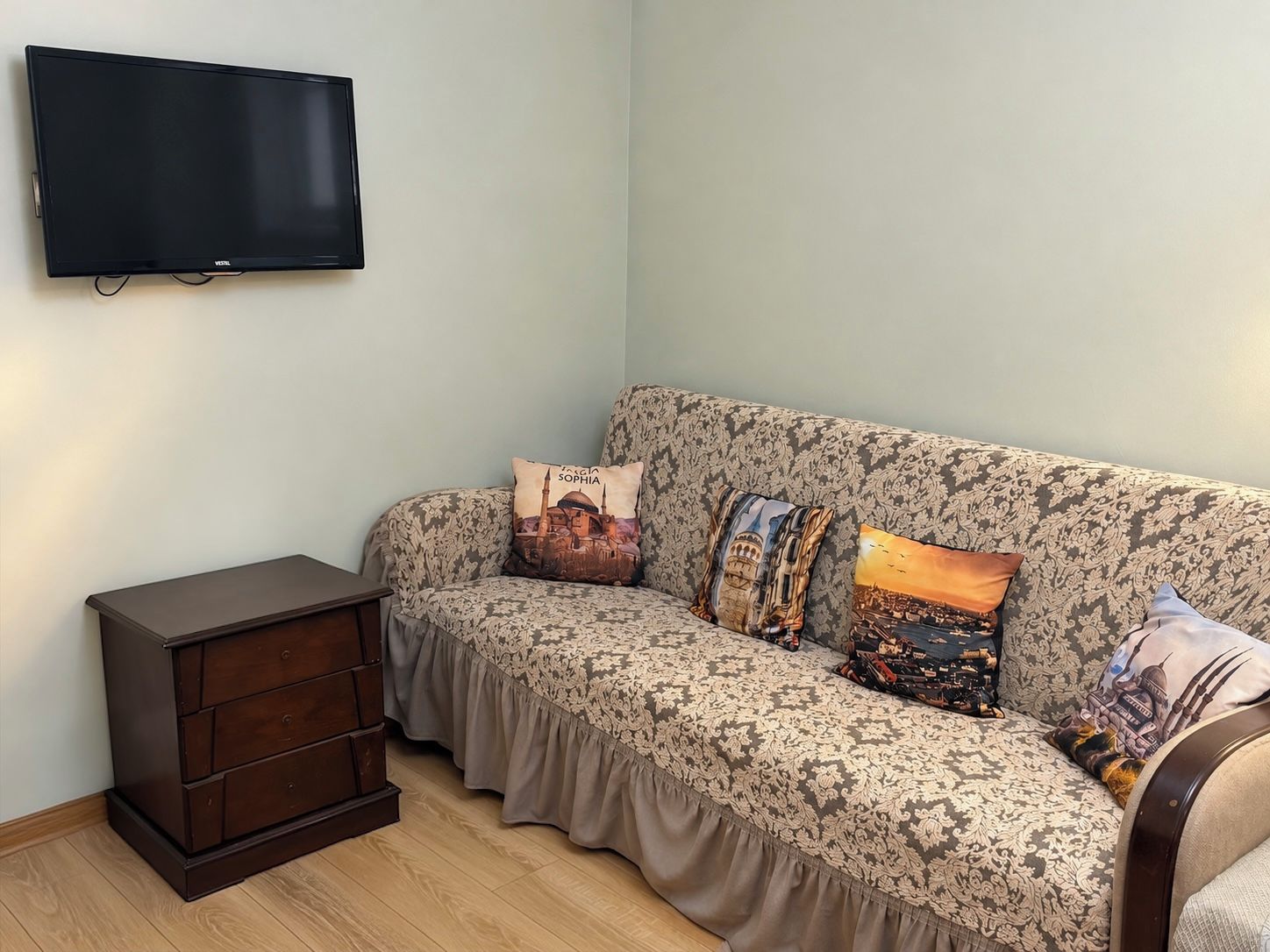 A living room with a sofa, a TV mounted on the wall, and a small wooden cabinet.