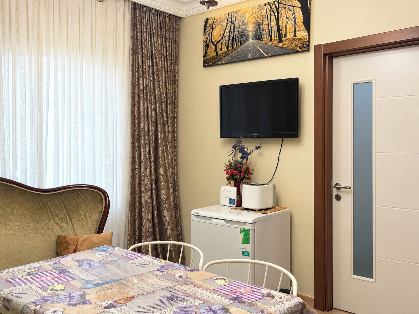 Habitación de hotel con cama, televisión montada en la pared, refrigerador pequeño y una puerta con panel de vidrio esmerilado.
