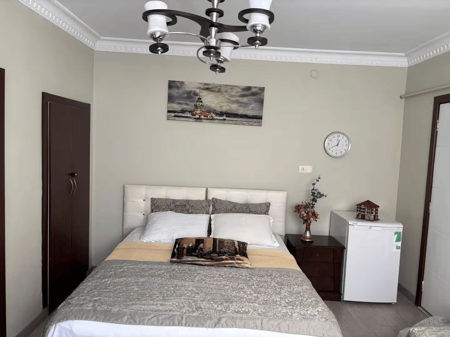 A bedroom with a bed, a clock, a small refrigerator, and a painting on the wall.