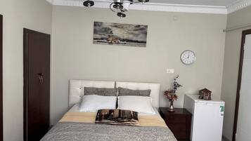 A bedroom with a bed, a clock, a small refrigerator, and a painting on the wall.