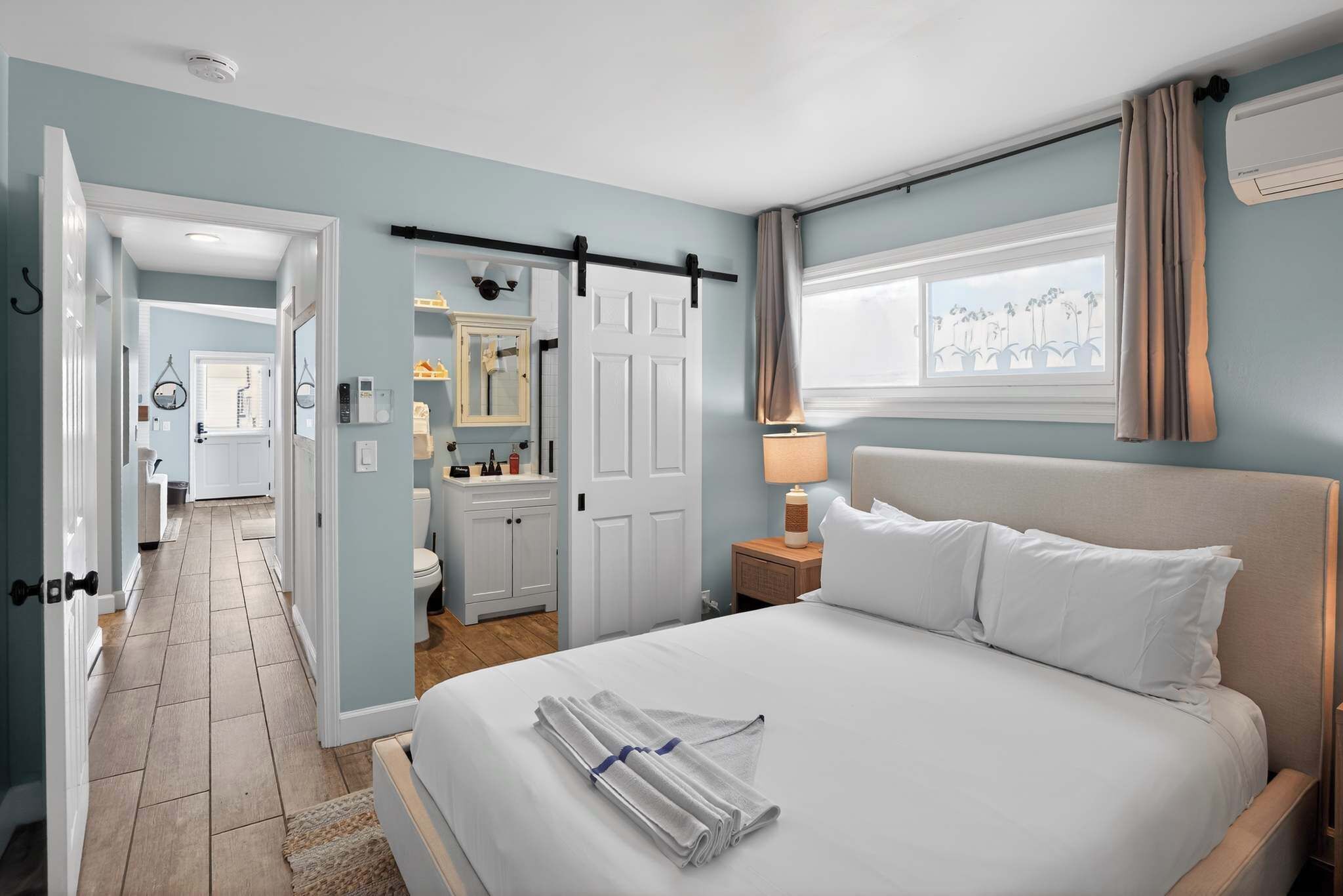 Habitación de hotel con cama, mesita de noche, ventana con cortinas, puerta y baño visible a través de una puerta abierta.